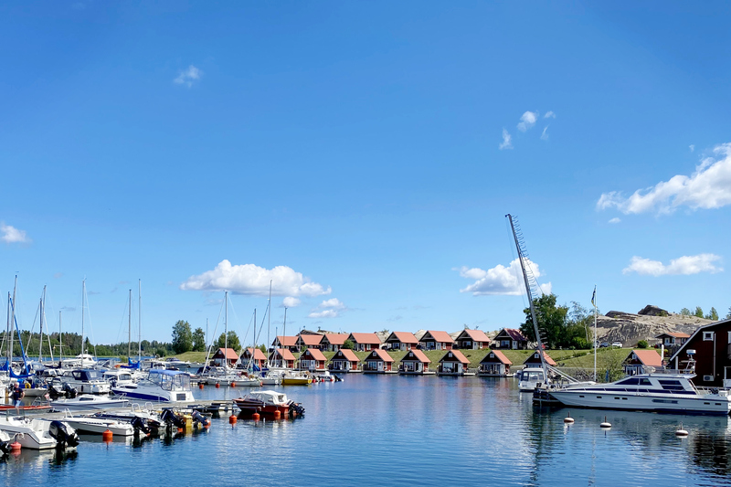 Bild på en massa röda stugor framför en hamn med segelbåtar. Blå himmel.