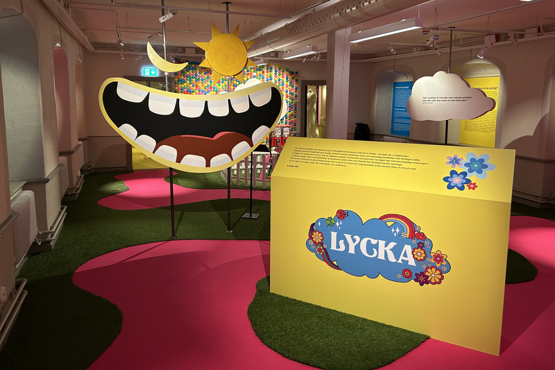 Bilden visar entren av utställning Lycka. En mun som skrattar, en sol och en måne. Golvet är rosa och grönt.