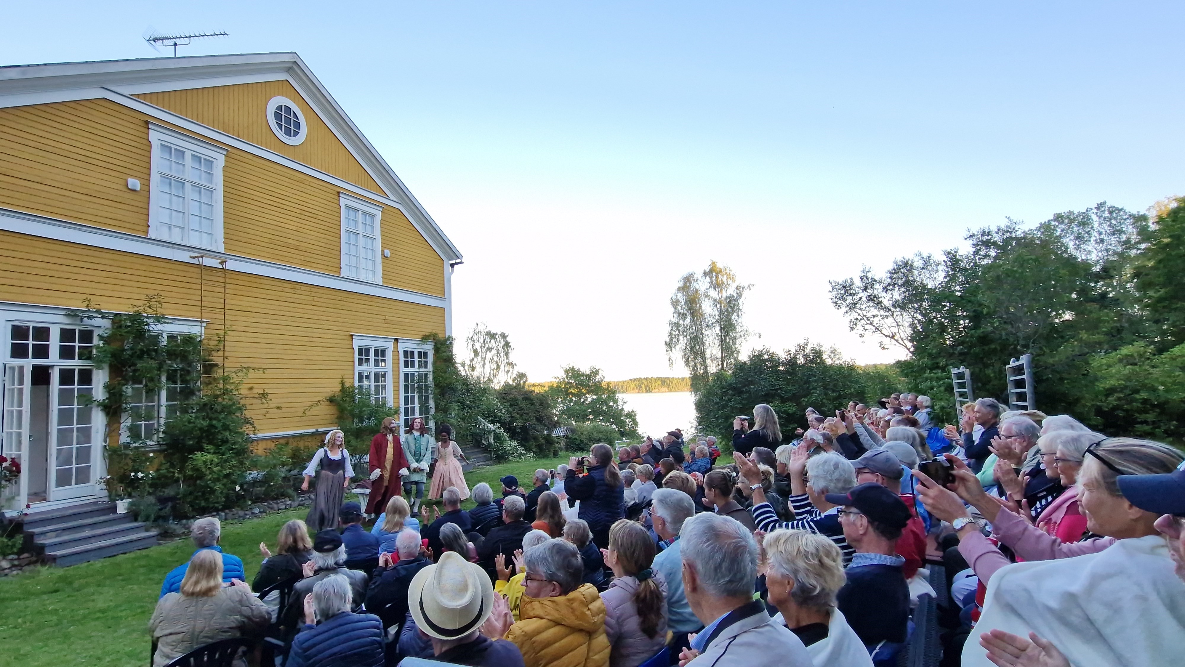 Bild på lycklig publik framför sekelskiftesvillan Gregersboda, med havsutsikt, blommande rosor och 4 1600- tals klädda skådespelare i ett applådtack.