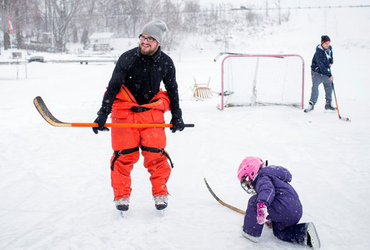 Pappa och dotter spelar hockey på is utomhus i vintrigt landskap.